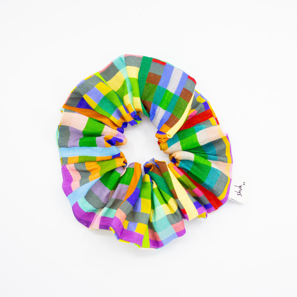Colourful scrunchie with a grid-inspired pattern featuring a mix of vibrant rectangles in green, yellow, red, purple, blue, and grey, designed to resemble a birds-eye view of patchwork farm fields, displayed in a circular shape on a white background.