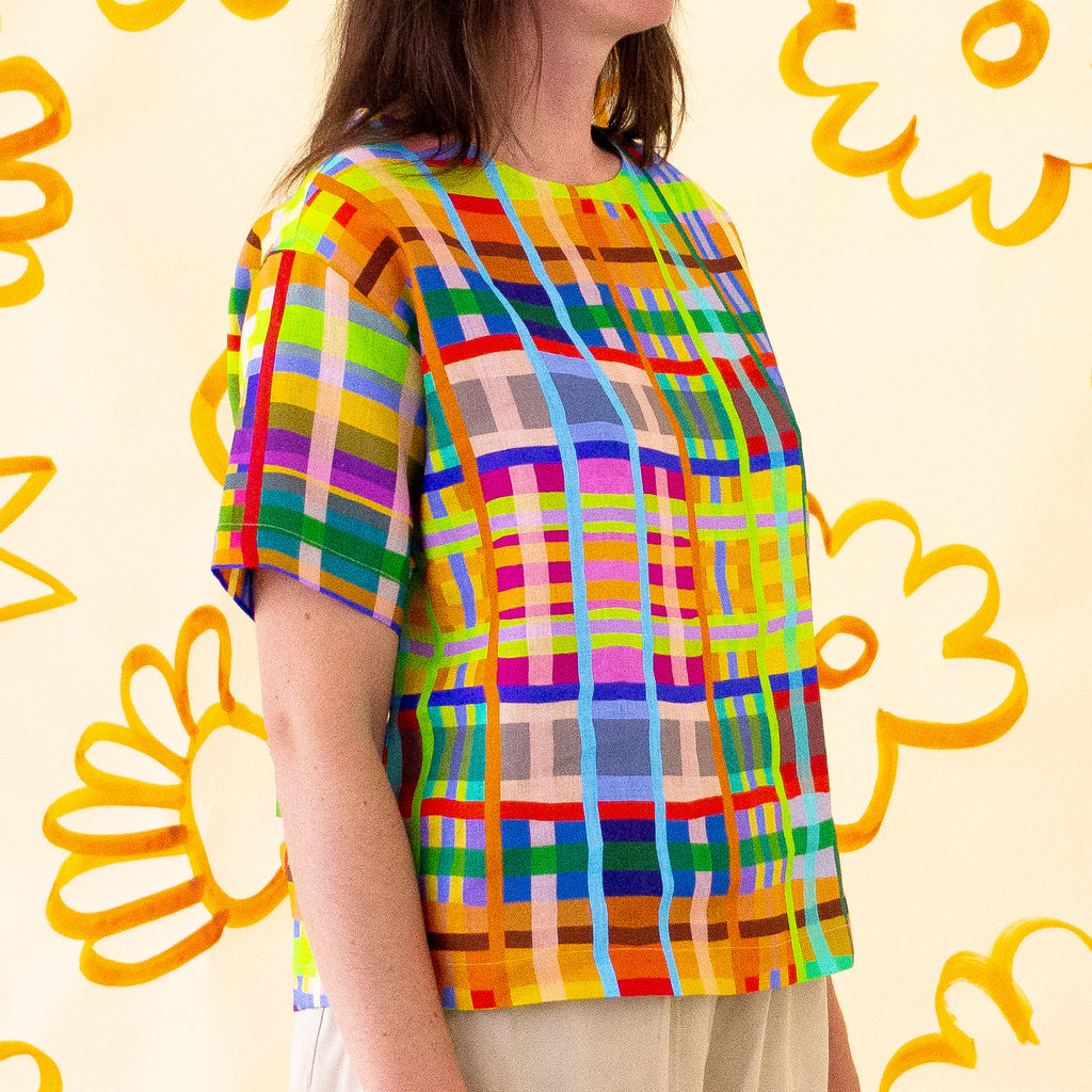 A side view of a model wearing the shuh Farm print linen top. Multicoloured lines in grid patterns resembling a birds eye view of a farm. 