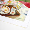 A watercolour art print depicting a breakfast scene with toast, a fried egg, a cup of tea, and a small bowl, shown on a white sheet with a floral tablecloth in the background.