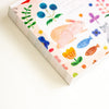A close-up of a notebook cover featuring a whimsical watercolour-style illustration of a pink bunny, blue berries, flowers, fish, and small animals on a warm cream background.
