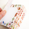 A close-up of a hand holding a fan of small decorative paper sheets with illustrated woodland motifs, including bears, berries, flowers, and leaves, on a white background.