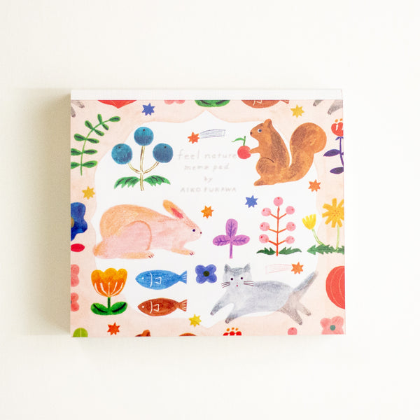 A square memo pad with a whimsical watercolour-style illustration pattern featuring a rabbit, squirrel, cat, flowers, and fish on a pale pink background, photographed against a clean white backdrop.
