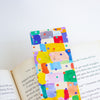 A colourful watercolour patchwork bookmark with multicoloured painted squares and dots, shown resting on open book pages against a white background.