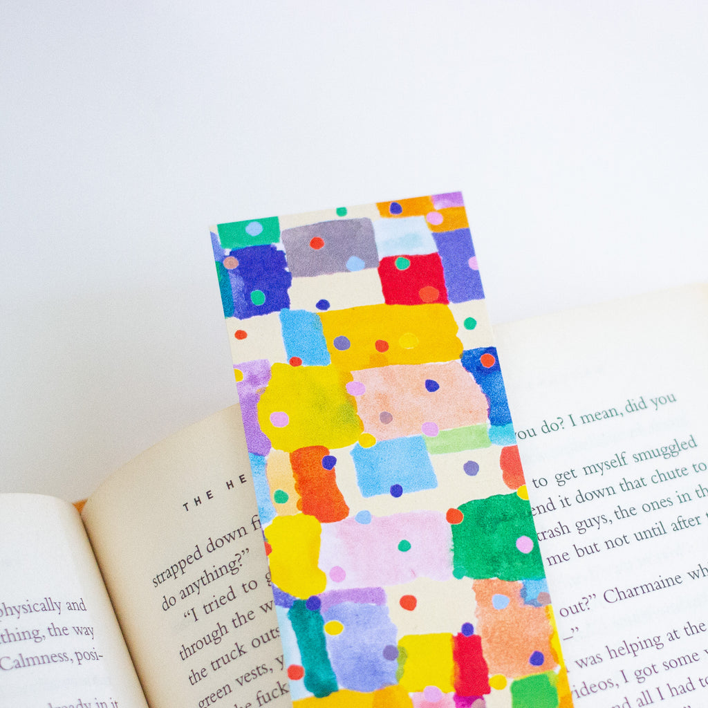 A colourful watercolour patchwork bookmark with multicoloured painted squares and dots, shown resting on open book pages against a white background.