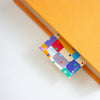 A close-up of a bright yellow-orange textured notebook or journal with a colourful patterned bookmark peeking out from the page. The bookmark features a patchwork design of blue, purple, red, green, and white squares with small polka dots.