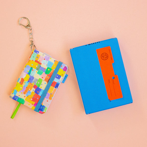 Colourful notebook with keychain and blue box with orange card on a peach background