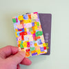 A hand holds a sticker with a colourful abstract watercolour pattern in front of a dark grey card against a pale mint green background.