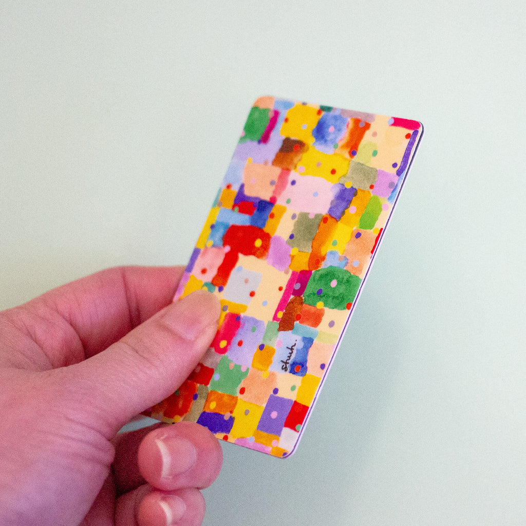 A hand holding a sticker with a colourful abstract patchwork pattern of painted blocks and dots on a pale mint background.