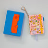 A pink patterned notebook keychain with a metal key ring and chain, shown next to a blue box with an orange card, on a light blue background.