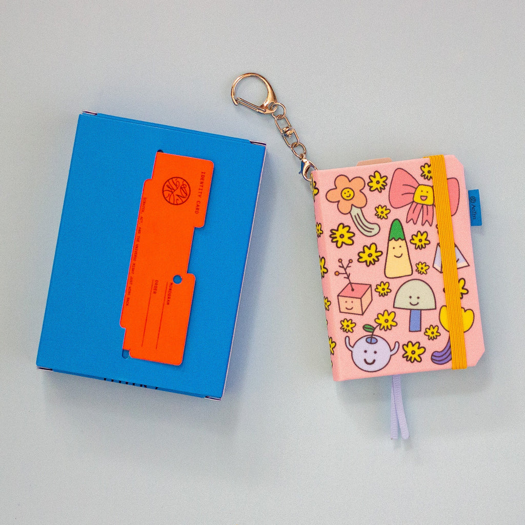 A pink patterned notebook keychain with a metal key ring and chain, shown next to a blue box with an orange card, on a light blue background.