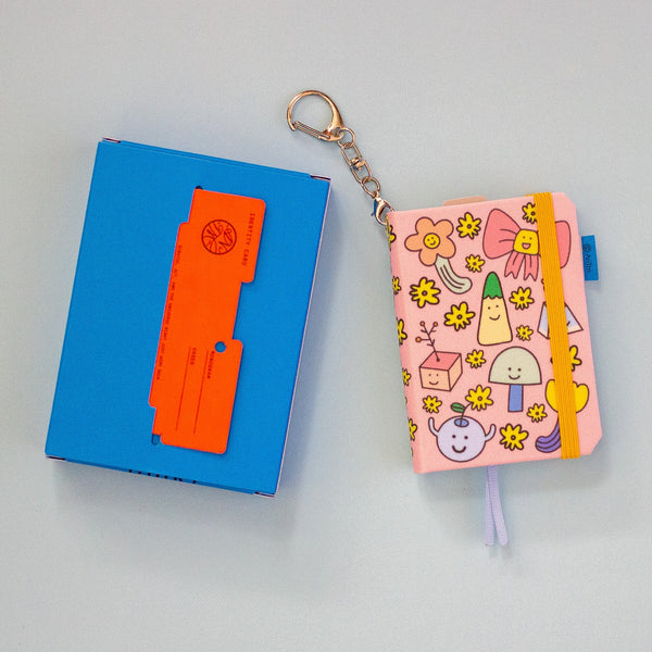 A pink patterned notebook keychain with a metal key ring and chain, shown next to a blue box with an orange card, on a light blue background.