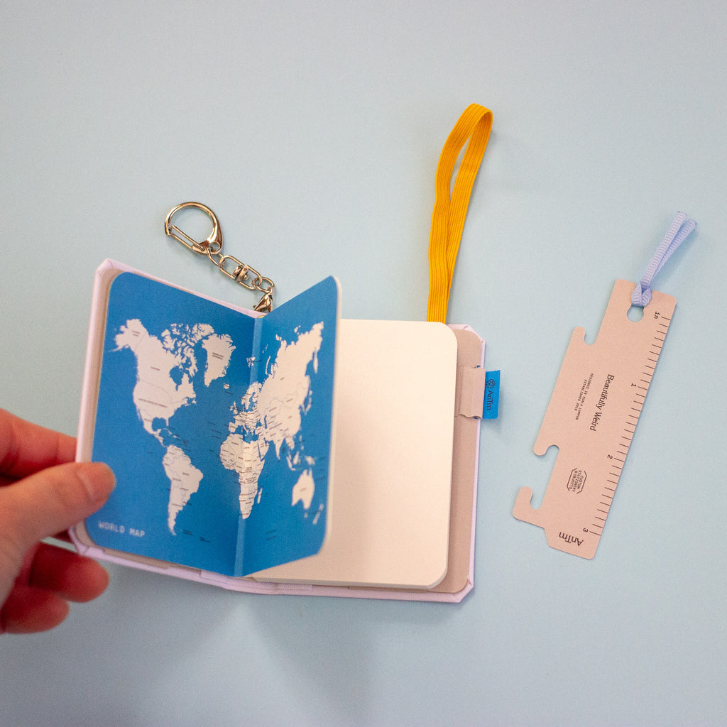 A hand holds an open notebook with a blue world map cover and a yellow elastic, next to a small ruler on a light blue background.