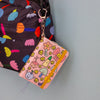 A notebook with pink fabric cover with cute cartoon characters and flower pattern, attached to a dark patterned bag with a silver key ring. 