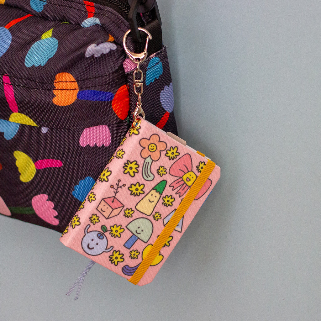 A notebook with pink fabric cover with cute cartoon characters and flower pattern, attached to a dark patterned bag with a silver key ring. 
