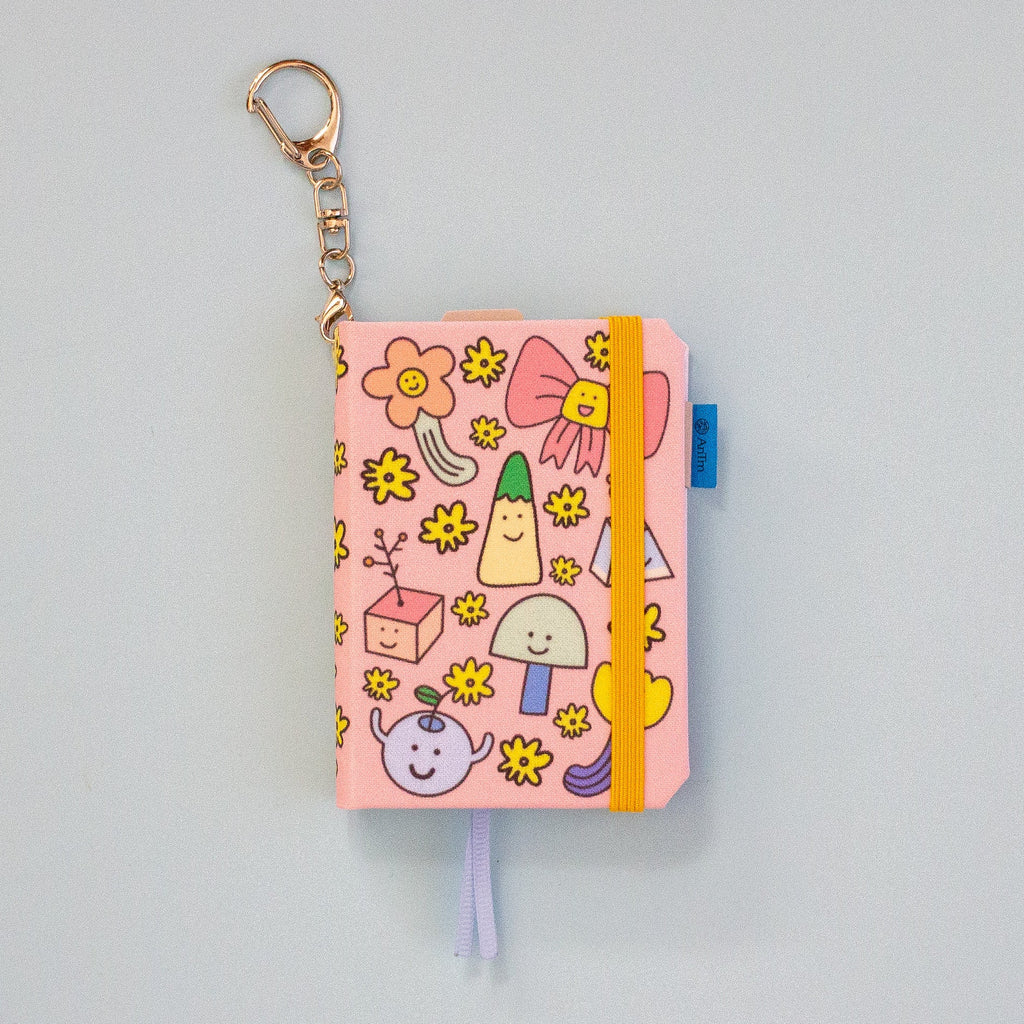 A pink mini notebook keychain with a gold keyring, decorated with cute cartoon illustrations. It features a mustard elastic closure band and a blue ribbon bookmark.