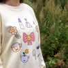 Beige sweatshirt with colourful cartoon-style illustrations of a flower, bow, cup, and more, worn by a person outdoors.