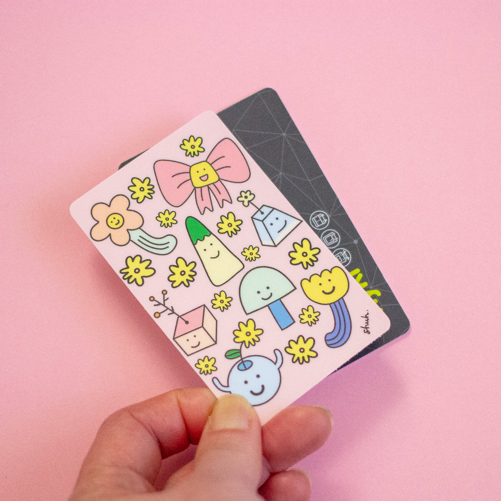 A hand holds a pink sticker sheet featuring cute cartoon stickers like a pink bow, yellow flowers, and a blue vase, with a dark grey card partially visible behind it on a solid pink background.