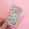 A hand holds a rectangular pink sticker featuring a cute pastel illustration of smiling flowers, mushrooms, a bow, and small doodles on a solid pink background.