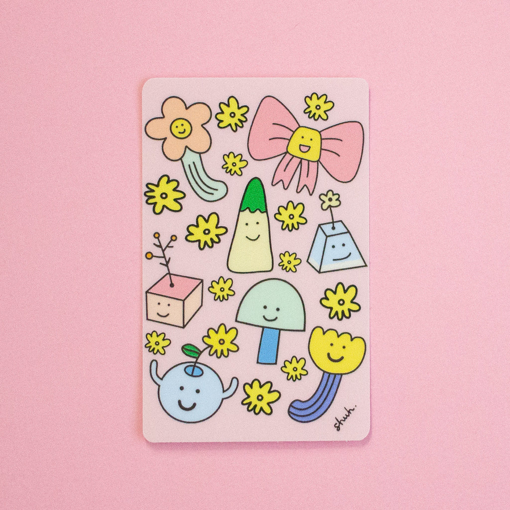A rectangular pink sticker featuring cute hand-drawn smiley-face illustrations, including yellow flowers, a pink bow, and small abstract shapes, photographed on a solid pink background.
