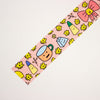 A strip of pink decorative tape featuring a playful pattern of yellow daisies and cute cartoon characters including a pink bow, a smiling mug, and small objects, laid diagonally on a plain white background.