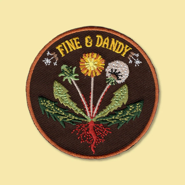A circular embroidered patch with the words 'FINE & DANDY' in yellow at the top, featuring a botanical design with green leaves, a yellow flower, a white flower, and red roots on a dark brown background, shown on a pale yellow backdrop.