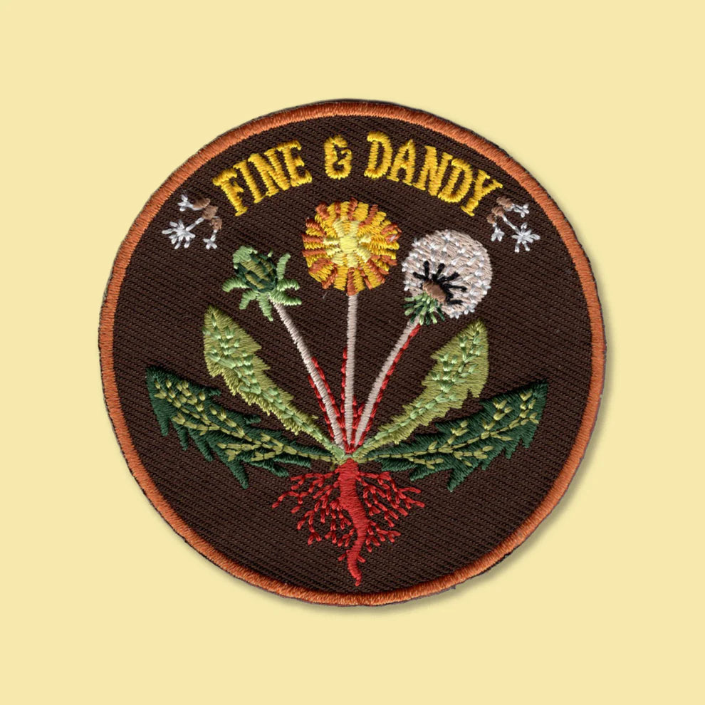A circular embroidered patch with the words 'FINE & DANDY' in yellow at the top, featuring a botanical design with green leaves, a yellow flower, a white flower, and red roots on a dark brown background, shown on a pale yellow backdrop.