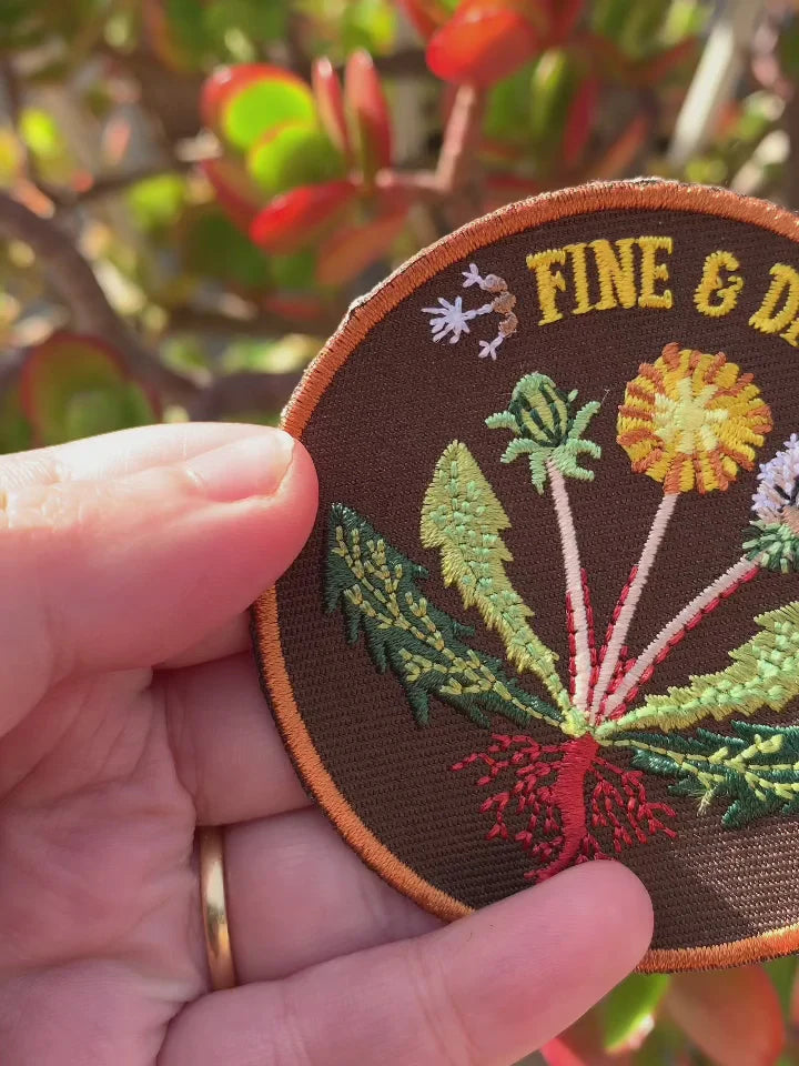 A close-up of a hand holding a round embroidered patch featuring a dandelion design with the text 'FINE & DANDY'.