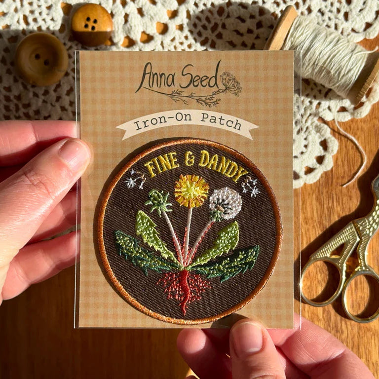 Iron-on patch with floral design held by a hand, on a wooden surface with sewing tools.