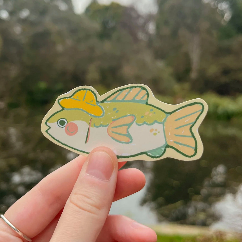 A hand holds a die-cut sticker shaped like a fish wearing a yellow hat, with a blurred outdoor background of trees and water.