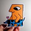 A hand holding a die-cut sticker featuring a stylised orange figure with one eye riding a blue spotted dog, with a small pink heart. The sticker has a visible artist credit reading 'Samantha Curcio' on the right edge.