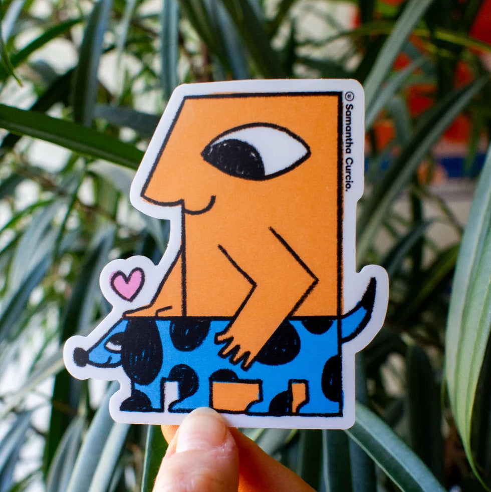 A hand holds a die-cut sticker featuring a stylised orange figure riding a blue spotted animal, with a small pink heart, in front of green plant leaves.