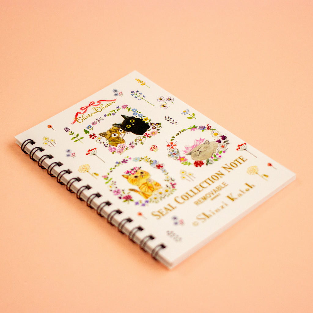 A spiral-bound notebook titled 'SEAL COLLECTION NOTE' by Shinzi Katoh featuring cute illustrated cats and floral wreaths on a cream cover, photographed on a solid peach background.