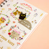 A close-up of a spiral-bound notebook with a white cover decorated with colourful cat illustrations, floral details, and a round sticker seal. The notebook is photographed on a soft peach-pink background.