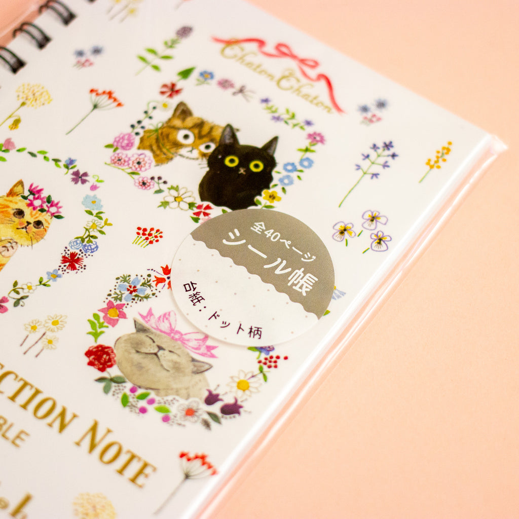 A close-up of a spiral-bound notebook with a white cover decorated with colourful cat illustrations, floral details, and a round sticker seal. The notebook is photographed on a soft peach-pink background.