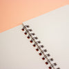 A close-up of an open spiral notebook with white dotted pages and a black wire binding, set against a peach-coloured background.