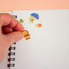 A hand is placing a small yellow character sticker with an orange outfit onto a white dotted notebook page, with other colourful stickers visible above it.