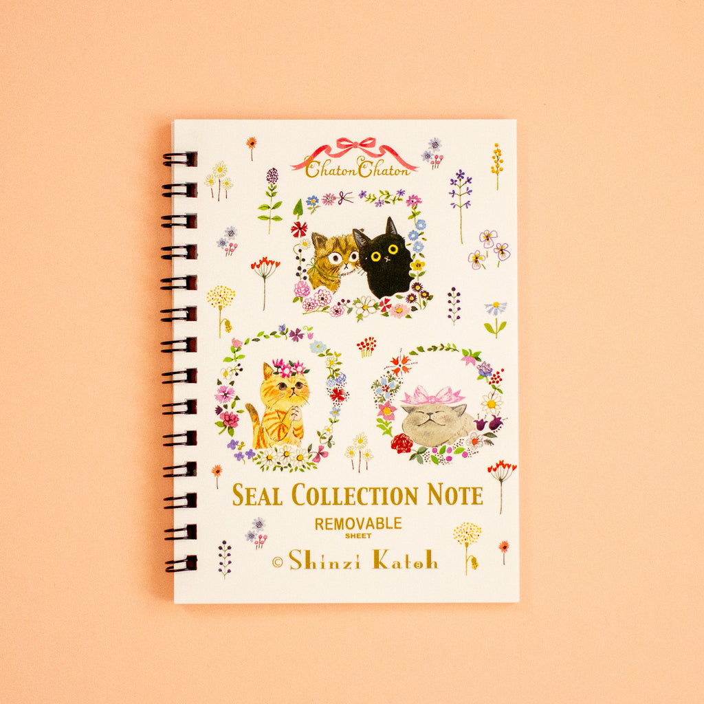 A spiral-bound notebook titled 'SEAL COLLECTION NOTE' with cute cat illustrations and floral designs on a cream cover, photographed against a peach background.