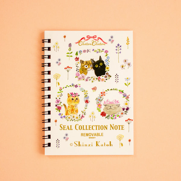 A spiral-bound notebook titled 'SEAL COLLECTION NOTE' with cute cat illustrations and floral designs on a cream cover, photographed against a peach background.