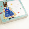 A square decorative memo pad with an illustrated rabbit in a blue dress on a light blue floral border, photographed at an angle on a white background.