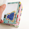 A hand holding a stack of colourful patterned pages on a white background. The top page features a whimsical illustration of a character in a blue dress on a light blue floral border, with other patterned cards fanned out underneath.