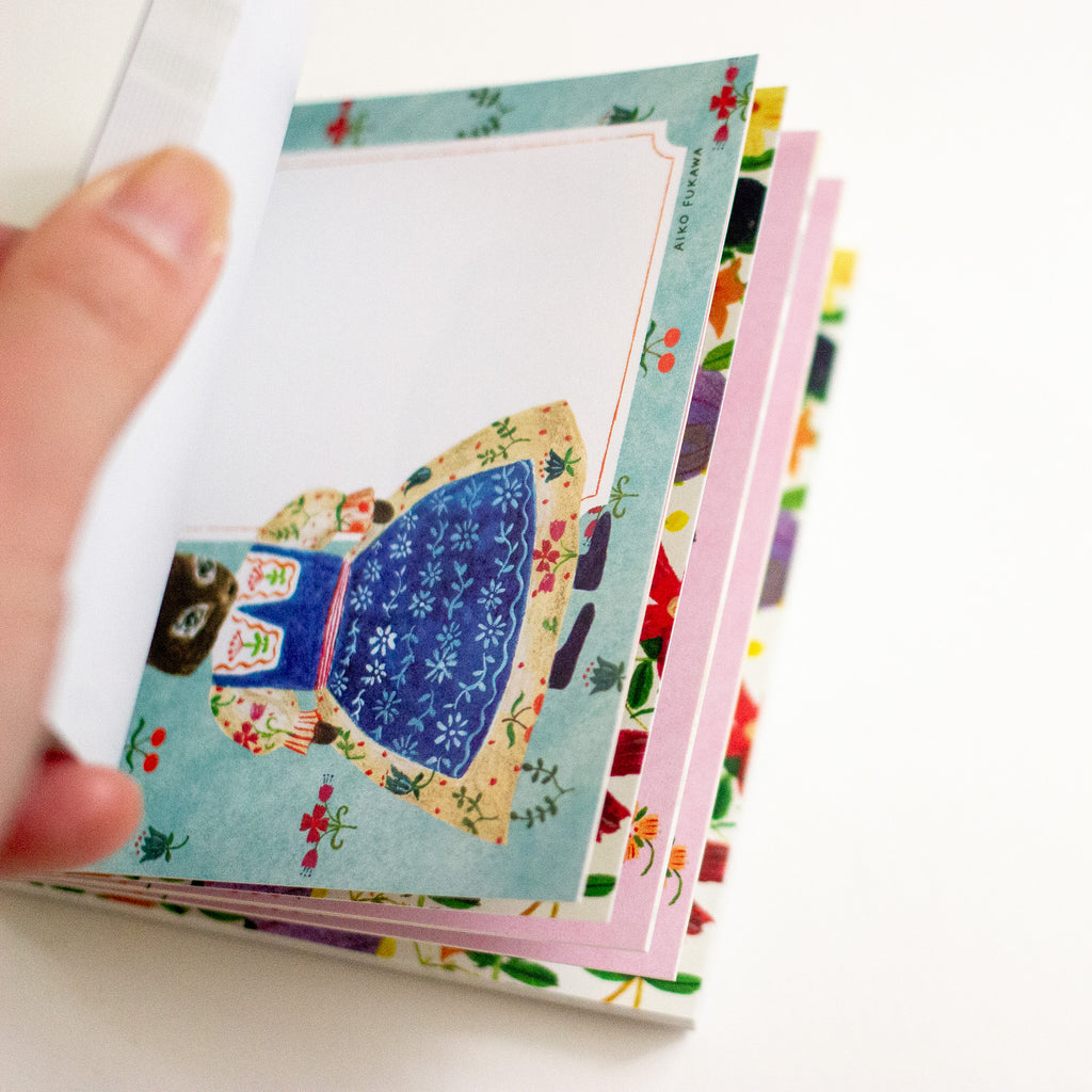 A hand holding a stack of colourful patterned pages on a white background. The top page features a whimsical illustration of a character in a blue dress on a light blue floral border, with other patterned cards fanned out underneath.