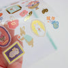 A hand holds a clear sheet of decorative stickers featuring cute bunny illustrations in pastel colours, including pink, yellow, blue, purple, and gold accents, against a bright white background.