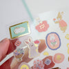 A hand holds a clear sheet of cute bunny-themed stickers against a bright white background. The stickers feature pastel-coloured rabbits, flowers, and decorative frames with gold foil accents.