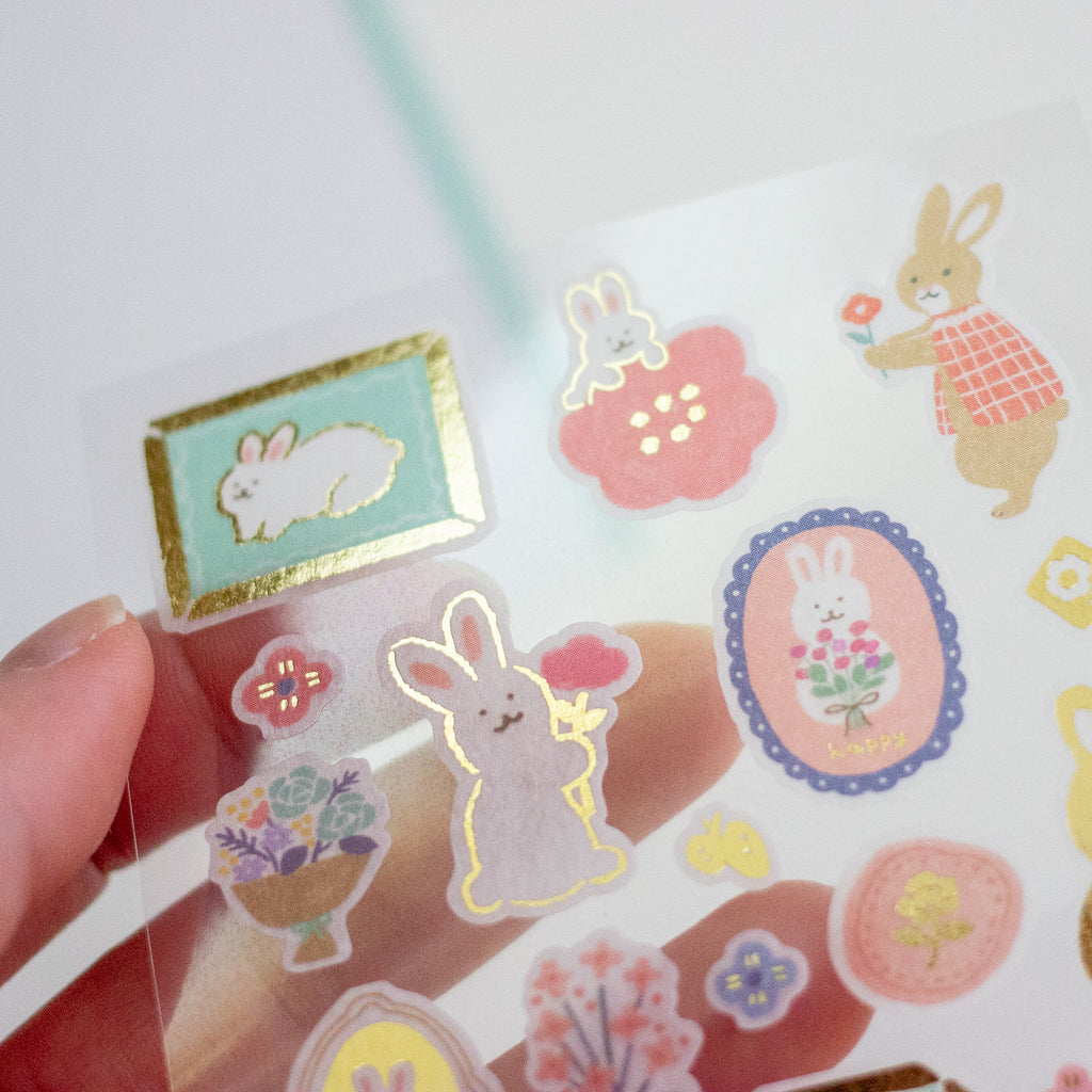 A hand holds a clear sheet of cute bunny-themed stickers against a bright white background. The stickers feature pastel-coloured rabbits, flowers, and decorative frames with gold foil accents.