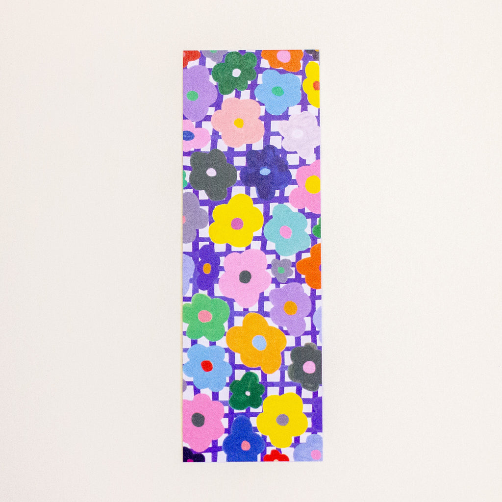 A tall, narrow rectangular bookmark featuring a dense, colourful floral pattern on a grid background, displayed flat against a light off-white backdrop.