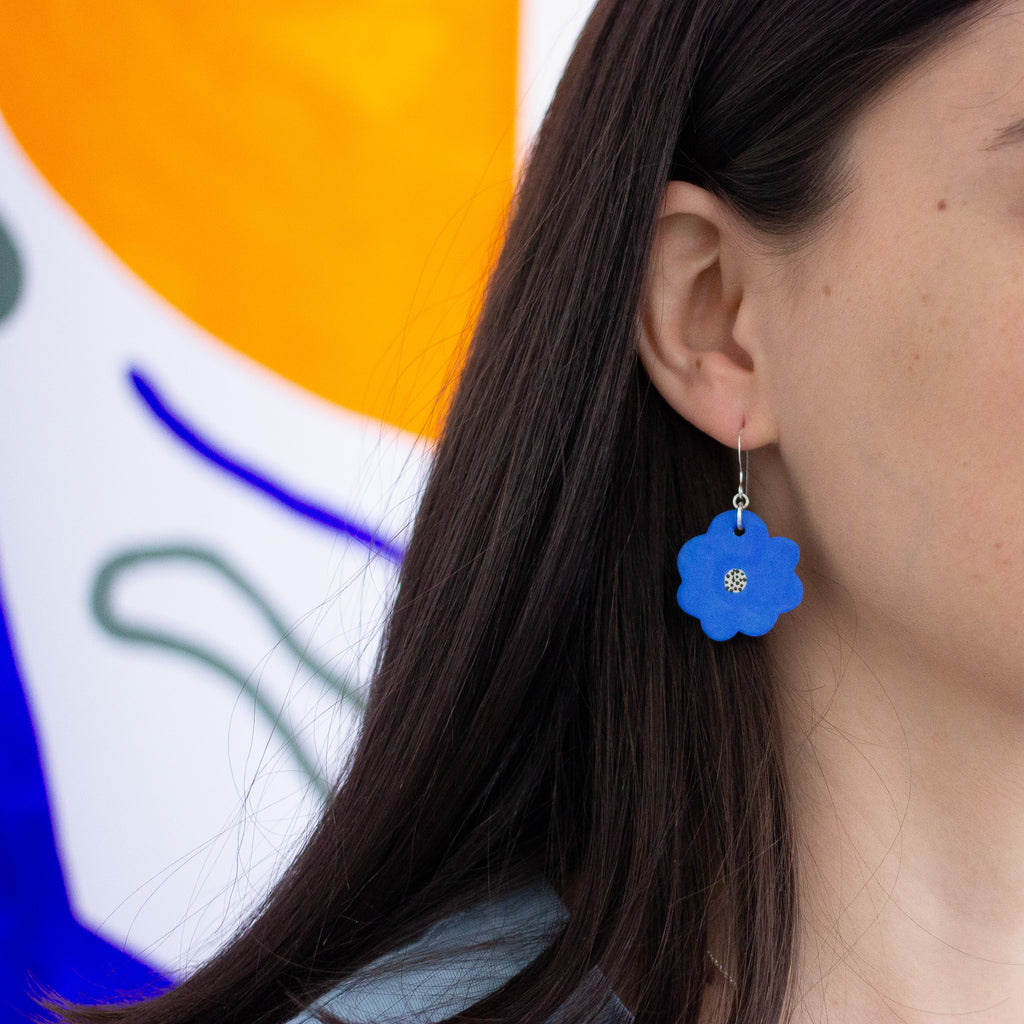 Close-up of a person wearing a bright blue handmade ceramic flower earring with a speckled centre. The earring dangles from a silver hook. A colourful abstract background is partially visible.