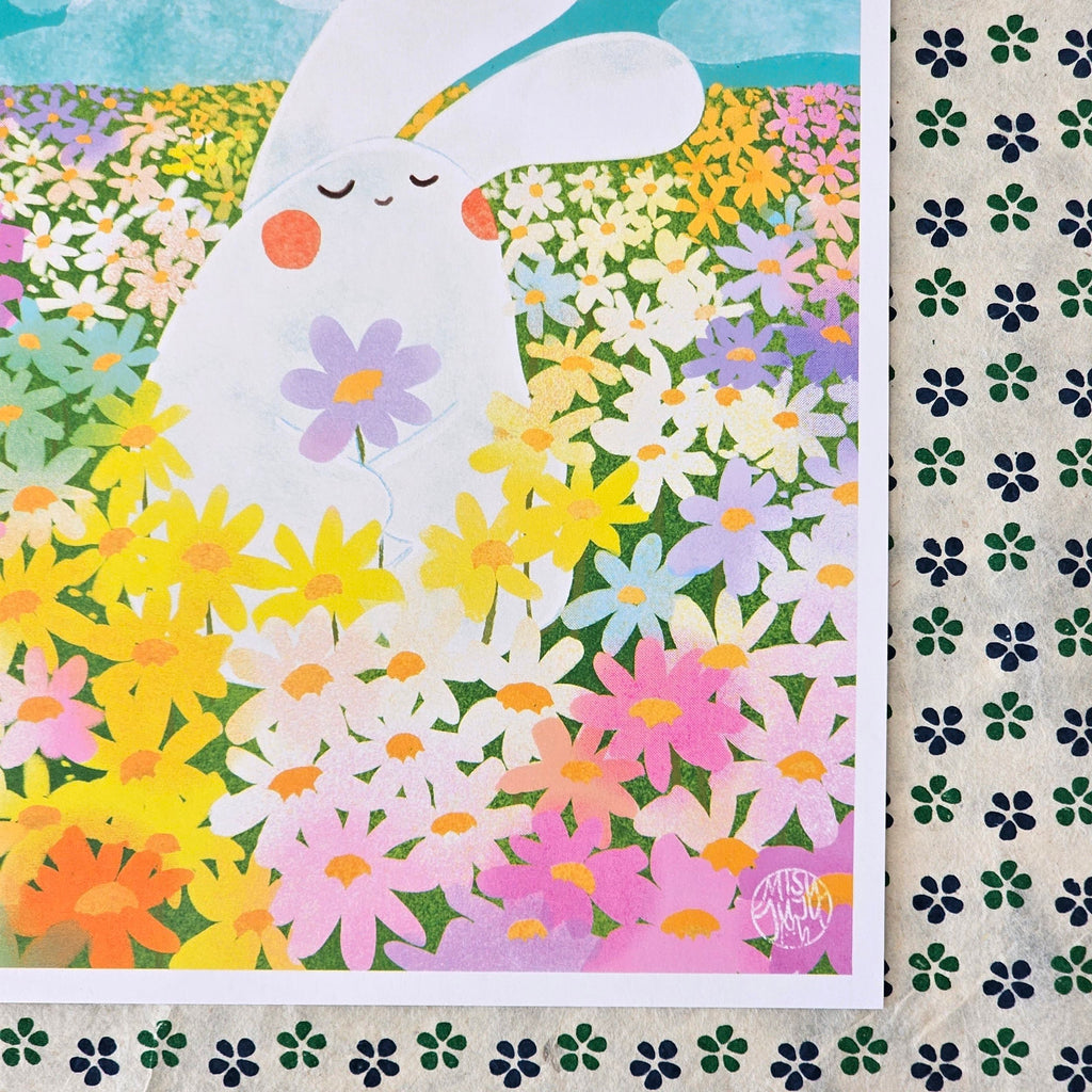 An art print featuring a cute white bunny surrounded by a colourful field of daisies, shown laid flat on patterned paper. The print has a white border and a small artist signature in the bottom right corner.