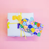 Closeup picture of the flower gift tag attached to a yellow string on a gift box. Print has hand drawn daisies, over a blue and white chequered background. Flowers are multicoloured. 
