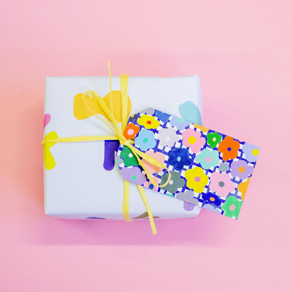 Closeup picture of the flower gift tag attached to a yellow string on a gift box. Print has hand drawn daisies, over a blue and white chequered background. Flowers are multicoloured. 