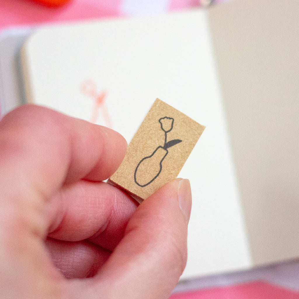 A hand holds a small rectangular stamp with a simple black line drawing of a flower in a vase, with a blurred notebook in the background.